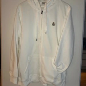 Vit hoodie från Moncler med dragkedja - Säljer en clean vit hoodie från Moncler med dragkedja framtill, huva med snörning och klassisk logga på bröstet. Tröjan har känguruficka och är tillverkad i mjuk bomull. Perfekt för dig som gillar stilrena och exklusiva plagg.