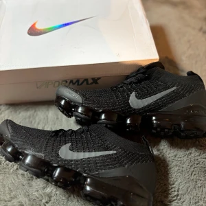 Nike Vapormax Flyknit svarta sneakers - Svarta Nike Vapormax Flyknit sneakers med stickad ovandel och reflekterande Swoosh på sidorna. Skorna har en unik, genomskinlig platt sula med synliga luftkuddar och snörning framtill. Perfekt för dig som gillar sportig och modern stil.