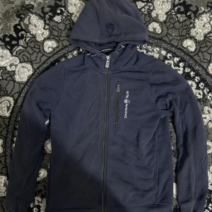 Sail Racing Marinblå hoodie  - Säljer en marinblå hoodie från Sail Racing med dragkedja framtill och en extra dragkedjeförsedd bröstficka. Tröjan har huva med broderad logga och snygga detaljer. Perfekt för dig som gillar sportig och stilren look.