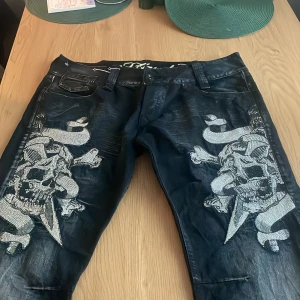 Svarta Ed Hardy jeans med döskallemotiv - Unika svarta jeans från Ed Hardy med stora döskalle- och benmotiv i silverfärgade stenar på båda benen. Klassisk femficksmodell med tydlig Ed Hardy-logga och läderpatch bak. Snyggt glansig finish och detaljer som sticker ut.