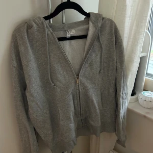 Grå hoodie med dragkedja från H&M - Mysig grå hoodie från H&M i storlek XS. Tröjan har huva med snörning, dragkedja framtill och två stora fickor. Perfekt för chill dagar och enkel att matcha med jeans eller mjukisbyxor.