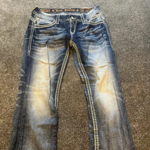 Rock Revival blå jeans med broderade fickor - Snygga blå jeans från Rock Revival med kraftiga kontrastsömmar och broderade bakfickor med nitar. Jeansen har en faded effekt och raka ben, klassisk femficksdesign och detaljerade sömmar längs benen. Perfekt för dig som gillar statement-jeans.