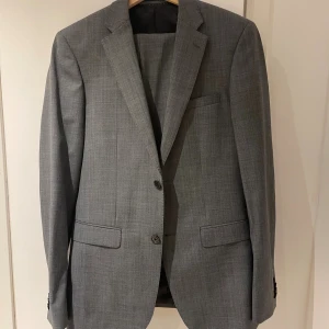 Grå rutig kostymkavaj och byxa - Klassisk grå kostym med diskret rutigt mönster, består av kavaj med två knappar och matchande kostymbyxa. Kavajen har fickor med lock och smal krage, byxan har bälteshällor och bakficka. Perfekt för dig som gillar stilren och tidlös look.