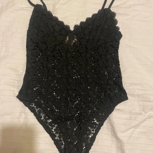 Svart spetsig body med smala band - Säljer en svart body med smala axelband och v-ringad hals. Toppen är helt täckt av virkad spets med blommönster som ger en snygg och unik look. Perfekt att styla med jeans eller kjol för en trendig outfit.