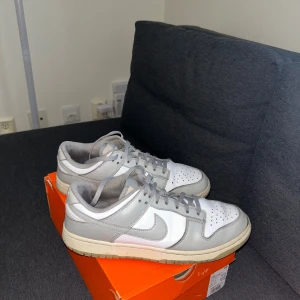 Nike grey frogs i grått och vitt (nypris 1499kr) - Nike Dunk Low sneakers i stilren grå och vit färg. Skorna har klassisk snörning, rund tå och platt sula. Ovandelen är i skinn med perforerade detaljer på tån för extra ventilation. Snygg kontrast mellan de grå panelerna och den vita basen, perfekt för dig som gillar minimalistisk stil.
