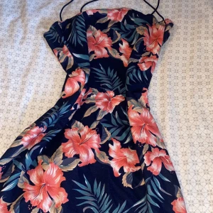 Blommig marinblå kortklänning - Supersnygg marinblå kortklänning med tropiskt blommigt mönster i rosa och beige toner. Klänningen har smala axelband och figurnära passform som framhäver siluetten. Perfekt för sommarens alla tillfällen när du vill sticka ut med färg och mönster.