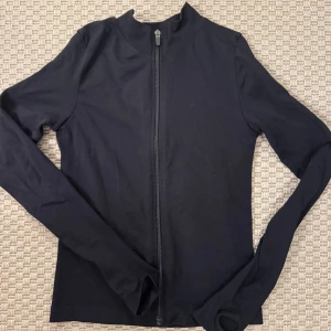 Svart långärmad ziptröja - En svart långärmad tröja med hel dragkedja framtill och hög krage. Tröjan är figurnära och har tumhål vid ärmsluten, vilket gör den perfekt för lager-på-lager eller till träning.