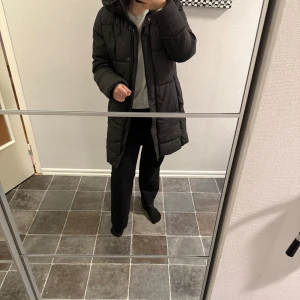 Svart lång pufferjacka med huva - Säljer en svart lång pufferjacka med huva och dragkedja framtill. Jackan har en quiltad design, dragsko i huvan och praktiska fickor på sidorna. Perfekt för kalla dagar när du vill hålla dig varm och bekväm. Flitigt använd men är i mycket bra skick! 