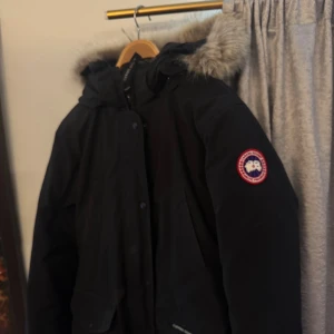 Svart Canada Goose parkas med päls - Säljer en svart parkas från Canada Goose med klassisk patch på ärmen och stor huva med fluffig päls. Jackan har flera fickor, dragkedja och knappar framtill. Perfekt för kalla dagar och riktigt snygg streetstil. Höst vibe