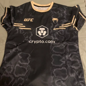 Svart UFC x Venum  - Snygg svart  från Venum i samarbete med UFC. Tröjan har guldiga detaljer, tryck med crypto.com och coolt mönster som påminner om camouflage och hexagoner. Tillverkad i lätt och ventilerande mesh, perfekt för träning..