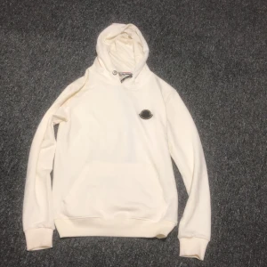 Vit hoodie från Moncler med logga - Säljer en stilren vit hoodie från Moncler med svart och gul Moncler-logga på bröstet. Tröjan har huva, känguruficka och ribbade muddar. Perfekt för dig som gillar exklusiva streetwear-plagg med clean look.