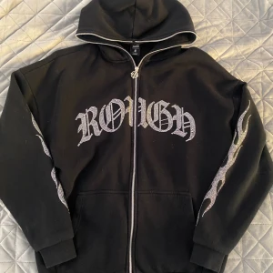 Svart hoodie från Black Squad med stenar - Cool svart hoodie från Black Squad i oversized fit. Stora silvriga stenar bildar texten 'ROUGH' på bröstet och flames längs ärmarna. Dragkedja framtill och huva. Perfekt statement-plagg för dig som gillar streetstyle.