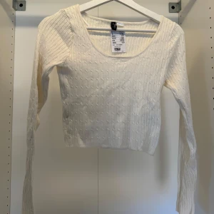 Vit ribbad långärmad topp från H&M - Säljer en fräsch vit långärmad topp från H&M med ribbad struktur och diskret mönster. Toppen har en rundad halsringning och är croppad i modellen, vilket ger en modern vibe. Perfekt att styla med jeans eller kjol för en clean look.
