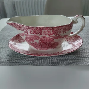 Enoch Wedgwood Woodland Gravy Boat med Fat - Vacker gravy boat med fat från Enoch Wedgwood, modell Woodland. Handgraverad dekor i rött med blommor och landskap. Tillverkad i England, mycket fint skick utan synliga skador eller slitage. Perfekt för samlare eller till festdukningen.
