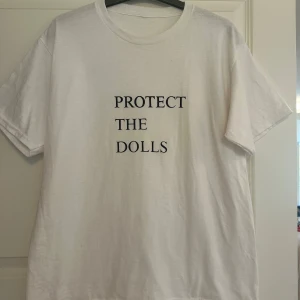 Vit t-shirt med texttryck - Vit t-shirt med svart text 'PROTECT THE DOLLS' på bröstet. Klassisk rund halsringning och korta ärmar. Enkel och clean design som passar perfekt till jeans eller shorts.