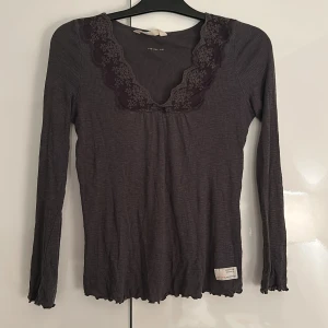 Mörkgrå ribbad topp med spets från Odd Molly - Säljer en mörkgrå långärmad topp från Odd Molly med ribbad struktur och v-ringad hals med spetskant. Små volangdetaljer vid ärmslut och nederkant samt en söt rosett framtill. Perfekt för dig som gillar detaljer och mjuka material, superskön och sparsamt använd, varit en favvo länge men passar inte längre tyvärr🥰många liknande tröjor säljs för mkt dyrare🥰