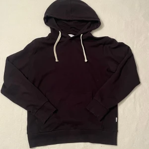 Svart hoodie från Jack & Jones - Svart hoodie från Jack & Jones med klassisk känguruficka och vita snören i huvan. Tröjan har lång ärm och ribbade muddar. Perfekt för chill dagar eller när du vill ha en enkel och stilren look. Stl: M