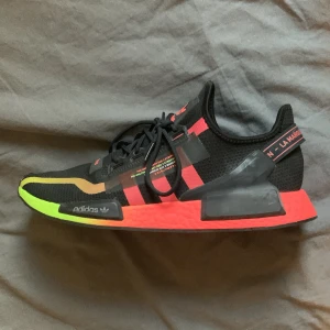 Adidas NMD V2 45 - Säljer ett par Adidas NMD sneakers med svart mesh-överdel, neonrosa och limegröna detaljer på sula och ovandel. Skon har klassiska tre ränder i rosa, snörning och platt sula. Perfekt för dig som gillar sportig och färgstark stil. KAN SÄLJA LÄNGRE PRIS FÖR SNABB AFFÄR