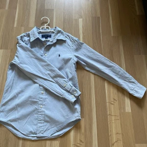 Ljusblå skjorta från Ralph Lauren -  ljusblå skjorta från Ralph Lauren med lång ärm och knappar framtill. Skjortan har den loggan på bröstet och är tillverkad i bomull. Perfekt för en clean och stilren look.