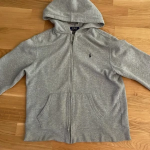 Grå hoodie från Polo Ralph Lauren XL - Klassisk grå hoodie från Polo Ralph Lauren i storlek XL. Tröjan har huva, dragkedja framtill och två stora fickor. Liten broderad logga på bröstet. Perfekt för chill dagar eller när du vill ha en enkel och stilren look.
