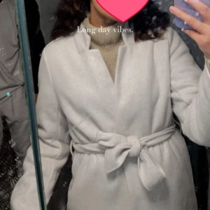 Ljusbeige kappa med bälte - Säljer en stilren ljusbeige kappa med knytbälte i midjan och klassisk krage. Jackan är långärmad och har en rak, elegant passform som ger en clean look. Perfekt för dig som gillar minimalistisk stil och vill ha en snygg jacka till höst och vinter. Jackan har en defekt då den fick ett lite hål, den har också blivit lite nopprig. Om bild skulle önskas så är det bara att säga till 