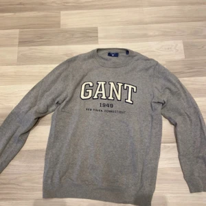 Grå stickad tröja från GANT - Snygg grå stickad tröja från GANT med broderad logga och texten 'GANT 1949 New Haven, Connecticut' på bröstet. Klassisk rund halsringning och långa ärmar. Perfekt för lager-på-lager och chill vardagsstil.