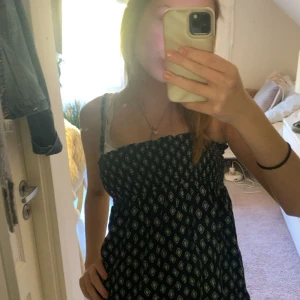 Svart kortklänning med mönster - Svart kortklänning med vita och beige geometriska mönster. Klänningen har smock upptill och tunna axelband. Materialet känns lätt och luftigt, perfekt för varma dagar. Passformen är normal och klänningen har en avslappnad siluett.