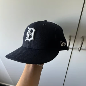 Svart New Era 59FIFTY keps Detroit - Säljer en svart New Era 59FIFTY keps med Detroit Tigers-logga i vitt framtill från hatstore. Klassisk flat brim och broderade detaljer. Kepsen är tillverkad i 100% polyester och har New Era-logga på sidan samt MLB-logga bak. 