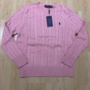 Rosa kabelstickad tröja Polo Ralph Lauren - Snygg rosa kabelstickad tröja från Polo Ralph Lauren med klassisk rund hals och broderad logga på bröstet. Tröjan har långa ärmar och breda ribbade muddar vid ärmslut och nederkant. Perfekt för dig som gillar preppy stil och vill ha något mjukt och bekvämt.