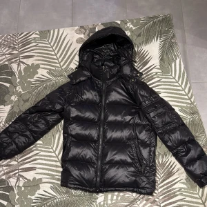 Svart pufferjacka med huva - Säljer en svart pufferjacka med huva och dragkedja framtill. Jackan har en glansig finish och en klassisk, väldigt bra passform som sitter på kroppen fint. Perfekt för kalla dagar när du vill hålla stilen enkel men ändå sticka ut. Hade bara en bild men om ni vill ha fler är de bara att skriva o fråga!