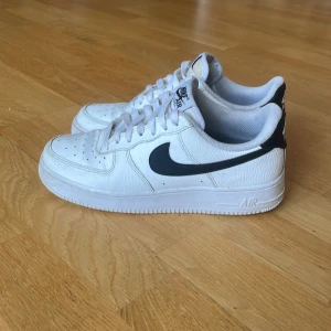 Nike Air Force 1 vita sneakers - Säljer ett par klassiska Nike Air Force 1 sneakers i vitt med svart swoosh och svart detalj på hälen. Inköpta på Zalando för ca 1500 kr. Skorna har snörning, perforerad tå och är tillverkade i skinn med platt sula. Perfekt för dig som gillar tidlös streetstil och vill ha en clean look.