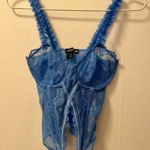 Blå spetskorsett från PrettyLittleThing - Säljer en blå korsett med spetsdetaljer och dekorativa axelband från PrettyLittleThing. Toppen har genomskinliga partier, vacker broderad spets och snörningsdetaljer framtill.