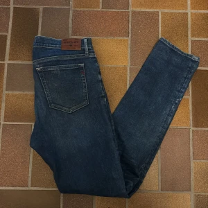 Replay jeans blå straight fit - Snygga blå jeans från Replay med klassisk femficksdesign och raka ben. Jeansen har en mörk tvätt med lätt slitning och detaljer som Replay-logga på bakfickan och läderpatch i midjan. Passar dig som gillar stilrena och tidlösa jeans. Jeansen är i nyshick och fickorna är lite små. Priset kan diskuteras 👍