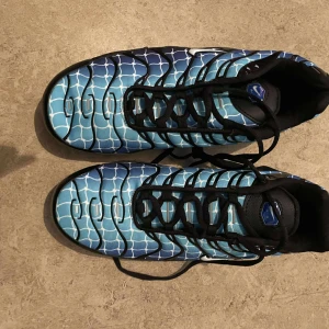Nike Air Max Plus blå mönstrade sneakers - Nike Air Max Plus sneakers med unikt blått och vitt mönster som påminner om vatten eller nät. Skorna har svarta detaljer, synlig Air Max-dämpning i sulan och svarta snören. Ovandelen är i syntetmaterial och mesh för en sportig vibe. Perfekta för dig som vill sticka ut.
