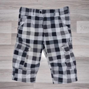 Rutiga cargoshorts i bomull - Snygga cargoshorts med stora fickor på sidorna och klassiskt rutigt mönster i svart, grått och vitt. Perfekta för sommaren med avslappnad passform och sköna att bära tack vare bomullsmaterialet. Stängs med dubbla knappar och har bälteshällor.