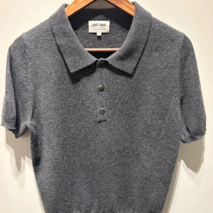 Grå kashmirpiké från Soft Goat - Mjuk och lyxig grå pikétröja i 100% kashmir från Soft Goat. Klassisk krage, tre knappar framtill och korta ärmar. Perfekt för dig som vill ha en stilren och bekväm look med premiumkänsla.