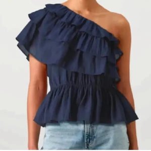 Marinblå volang topp oneshoulder - Säljer en marinblå topp med volanger och oneshoulder-design. Toppen har en snygg, asymmetrisk axel och är tillverkad i ett lätt bomullsmaterial. Den har en smickrande, rynkad midja och volangdetaljer som ger extra stilpoäng.
