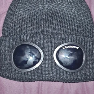 Svart C.P. Company mössa med goggles - Svart ribbstickad mössa från C.P. Company med ikoniska goggles framtill. Tillverkad i 100% bomull och har en cool, urban vibe. Perfekt för dig som vill sticka ut med streetwear-stil och gillar detaljer som syns.