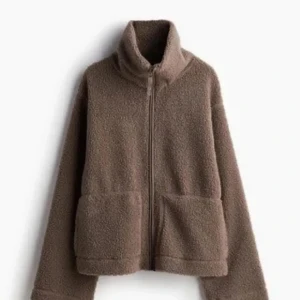 Brun teddyjacka med dragkedja - Mysig brun teddyjacka med hög krage och hel dragkedja framtill. Jackan har två stora fickor och är tillverkad i mjukt fleece-material. Perfekt för kyliga dagar och ger en avslappnad, trendig look.