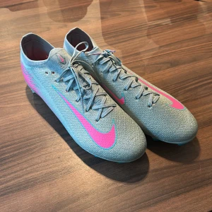 Nike Mercurial fotbollsskor blå/rosa - Säljer ett par Nike Mercurial fotbollsskor, de är blå och har rosa detaljer. Skorna har snörning, låg profil och en platt sula med dobbar för gräsplan. Ovandelen är i syntetmaterial med en stickad känsla och coolt mönster på sidan. Säljer pga för stor storlek då de är ca 27 cm.