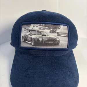 Mörkblå keps med bilmotiv - Snygg mörkblå keps med en stor patch framtill som visar ett svartvitt motiv av en klassisk sportbil. Kepsen har en mjuk, frottéliknande yta som ger en unik känsla och look. Perfekt för dig som gillar bilar och vill sticka ut med din stil.