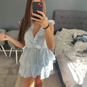 Ljusblå volangklänning från Nelly - Supersöt ljusblå kortklänning från Nelly med volanger, v-ringning och ärmlös design. Öppen rygg med snöre som man knyter. Helt ny aldrig använd. 