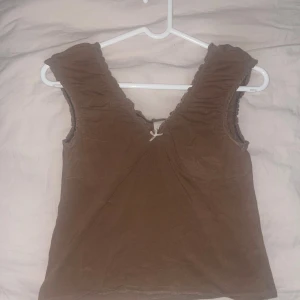 Brun linnetopp från Urban Outfitters - Supersöt brun linnetopp från Urban Outfitters med v-ringning och liten rosett framtill. Toppen är ärmlös med volangkant runt axlar och halsringning. Perfekt för dig som gillar en enkel men ändå söt stil.