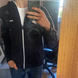 Svart hoodie från BOSS med vit dragkedja - Svart hoodie från BOSS i storlek S med vit dragkedja och vita detaljer längs ärmarna. Tröjan har huva med snörning och BOSS-logga på bröstet. Materialet är mjukt och känns som bomull, perfekt för chill dagar. Snygg och enkel design med normal passform. Ny pris ca 1.300kr, mitt pris 600kr! Kan tänka mig prisförhandling vid snabb affär 