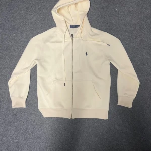 Polo Ralph Lauren Zip-Hoodie - Snygg beige hoodie från Polo Ralph Lauren med dragkedja och klassisk blå logga på bröstet. Tröjan har huva med snörning, två fickor fram och ribbade muddar. Perfekt för chill dagar och enkel att matcha med jeans eller joggers.