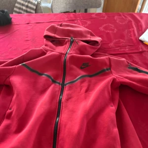 Röd hoodie från Nike med dragkedja - Snygg röd hoodie från Nike med hel dragkedja framtill och svart Nike-logga på bröstet. Tröjan har huva, långa ärmar och en cool svart detaljlinje över bröstet. Perfekt för dig som gillar sportig och avslappnad stil. Storlek S