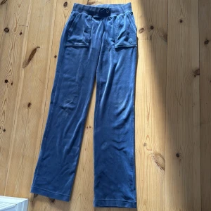 Blå velourbyxor Juicy Couture XS - Säljer ett par blå mjukisbyxor från Juicy Couture i velour med bred resår i midjan och två stora fickor fram med knappdetaljer. Byxorna har en rak och loose passform, perfekt för chill dagar. Ikonisk Juicy Couture-logga broderad på ena fickan. Tröjan i samma färg passar superbra tillsammans