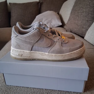 Nike Air Force 1 i ljusgrå mocka - Nike Air Force 1 sneakers i ljusgrå mocka och mesh. Klassisk låg modell med rund tå, snörning och gulddetaljer på snörningen samt logga på plösen. Snygg och stilren design med platt sula och diskret Nike Swoosh på sidan.