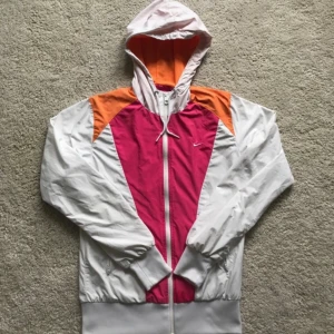 Nike färgglad vindjacka med huva - Nike vindjacka i vitt, rosa och orange med huva och hel dragkedja. Jackan har coola färgblock, meshfoder och snörning i huvan. Perfekt för dig som gillar sportig och retro stil. Tillverkad i 100% polyester.