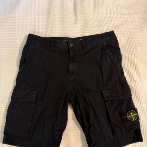 Svarta cargo shorts från Stone Island - Svarta cargo shorts från Stone Island med klassisk logopatch på benet. Modellen har flera fickor med lock och knappstängning, samt bälteshällor i midjan. Tillverkade i mjuk bomull för skön känsla och snygg streetstil. Perfekta för sommaren och chill dagar. Nypris ligger på 3500kr